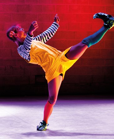 Ein Kind in schwarz-weißem Ringelpullover, roten Leggings, gelbem Kleid, großen schwarzen Schuhen und roten Haaren tanzt auf der Bühne.