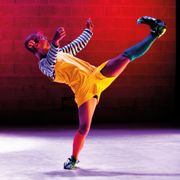 Ein Kind in schwarz-weißem Ringelpullover, roten Leggings, gelbem Kleid, großen schwarzen Schuhen und roten Haaren tanzt auf der Bühne.