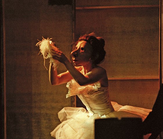 Eine Person mit einer weiblich gelesenen Maske kniet in einem Ballerinakleid und hält etwas hoch
