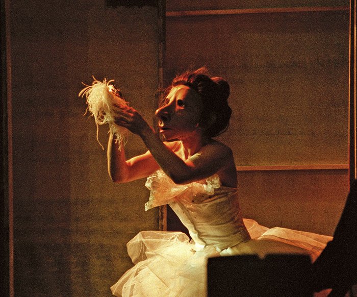 Eine Person mit einer weiblich gelesenen Maske kniet in einem Ballerinakleid und hält etwas hoch