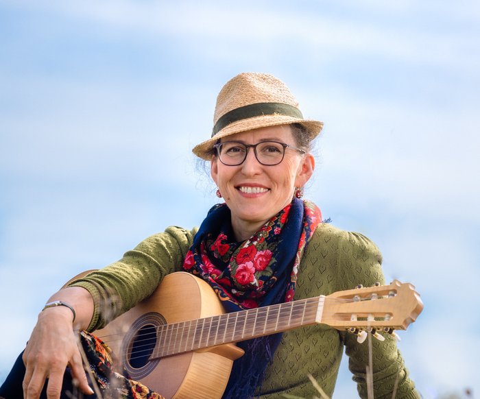 Portraitfoto Martina Schwarzman. Sie sitzt in einer Wiese und hat eine Gitarre vor sich.