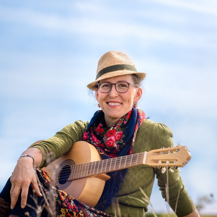 Portraitfoto Martina Schwarzman. Sie sitzt in einer Wiese und hat eine Gitarre vor sich.