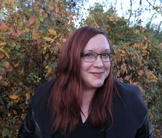 Porträt einer Frau mit rotem Haar und Brille im Freien, herbstliche Bäume im Hintergrund.