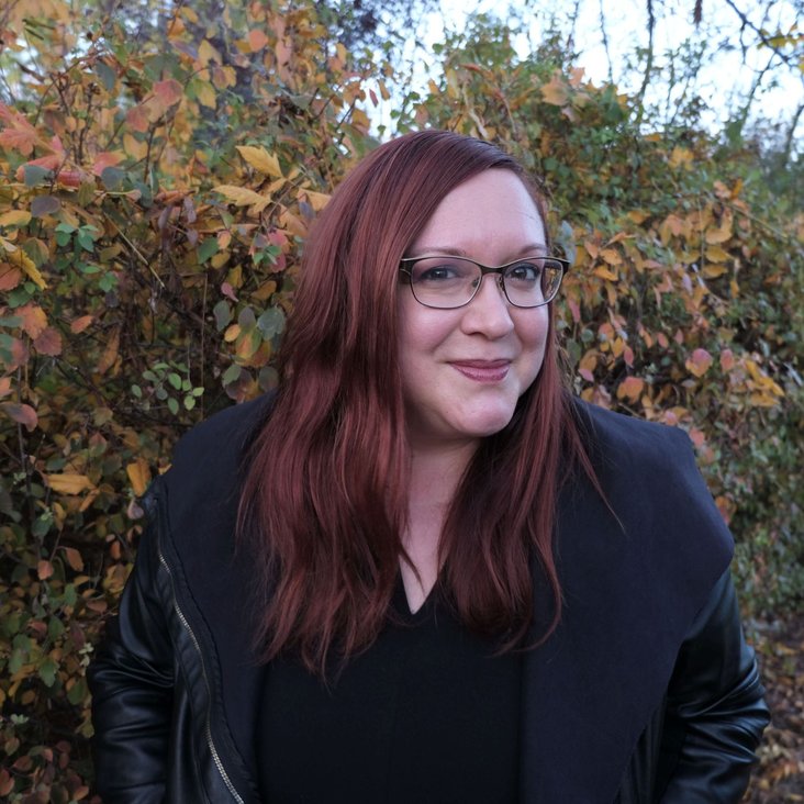 Porträt einer Frau mit rotem Haar und Brille im Freien, herbstliche Bäume im Hintergrund.