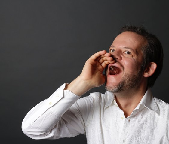 Porträt eines Mannes im weißen Hemd, der mit offener Hand ruft oder singt vor dunklem Hintergrund.