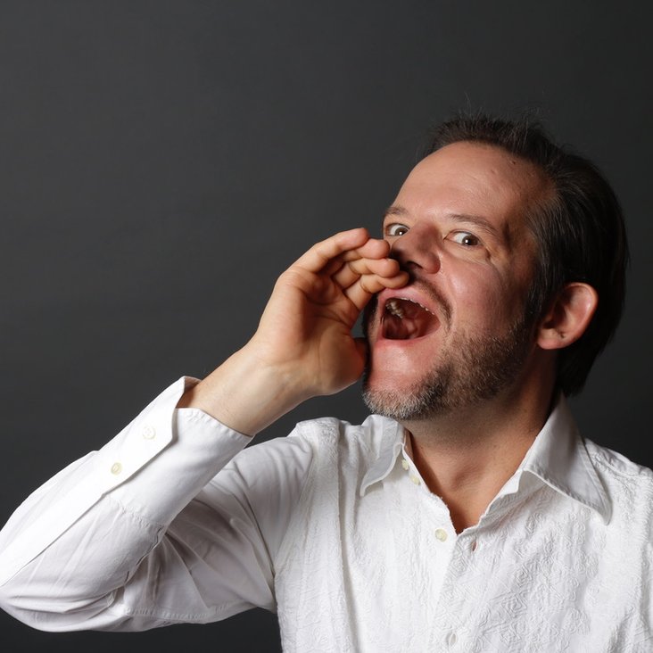 Porträt eines Mannes im weißen Hemd, der mit offener Hand ruft oder singt vor dunklem Hintergrund.