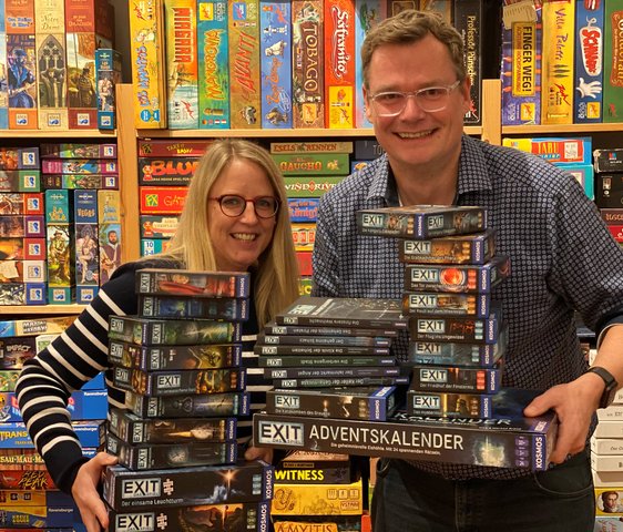 Zwei Personen stehen vor einem Regal voller Brettspiele, in ihren Händen halten sie zahlreiche EXIT Games Spielkartons und Special Editions.