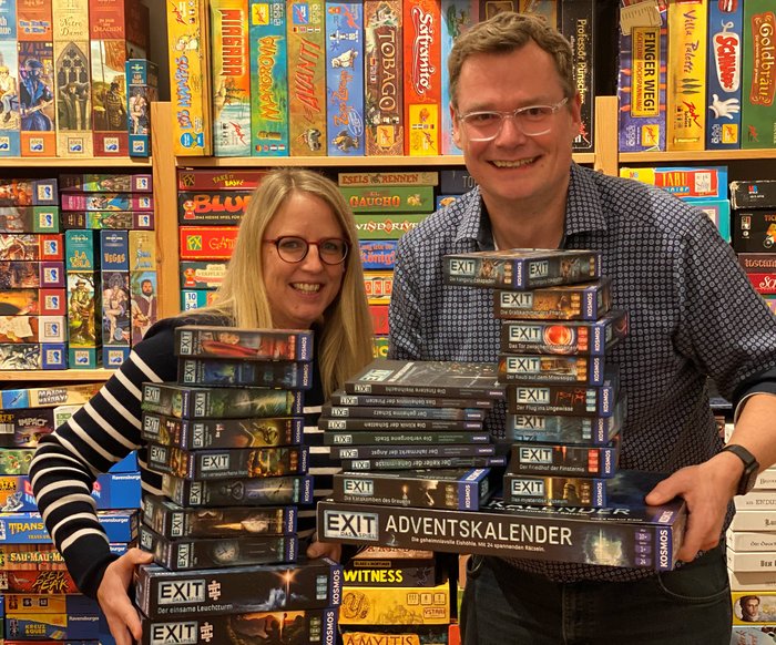 Zwei Personen stehen vor einem Regal voller Brettspiele, in ihren Händen halten sie zahlreiche EXIT Games Spielkartons und Special Editions.