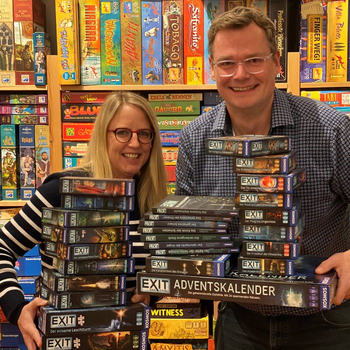 Zwei Personen stehen vor einem Regal voller Brettspiele, in ihren Händen halten sie zahlreiche EXIT Games Spielkartons und Special Editions.