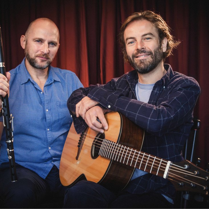 Die Musiker Brian Haitz & Brendan Mulholland sitzen vor einem roten Vorhang, sie haben ihre Instrumente in der Hand und lächeln in die Kamera