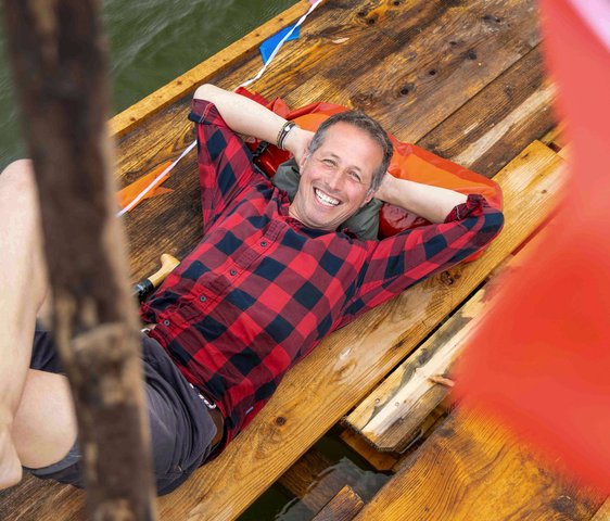 Portraitfoto von Willi Weitzel, er liegt mit dem Rücken auf einem Holzboot und trägt kurze Hosen und ein kariertes Hemd