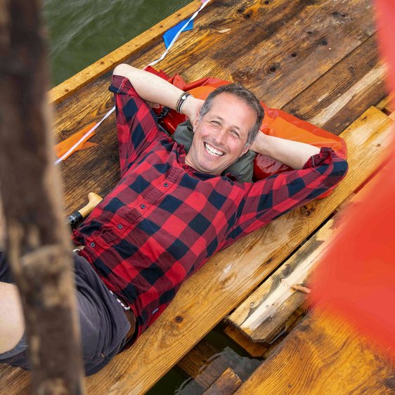 Portraitfoto von Willi Weitzel, er liegt mit dem Rücken auf einem Holzboot und trägt kurze Hosen und ein kariertes Hemd