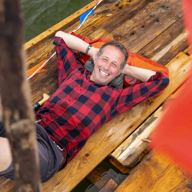 Portraitfoto von Willi Weitzel, er liegt mit dem Rücken auf einem Holzboot und trägt kurze Hosen und ein kariertes Hemd