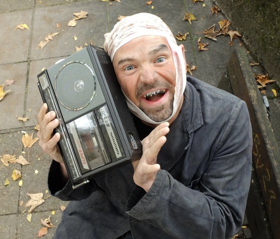 Ein Mann in schmutzigem Anzug, Kopfverband und schlechten Zähnen sitzt auf einer öffentlichen Parkbank und hat ein kleines Radio oder eine Jukebox in der Hand.