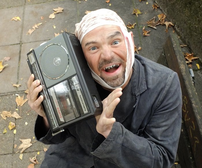 Ein Mann in schmutzigem Anzug, Kopfverband und schlechten Zähnen sitzt auf einer öffentlichen Parkbank und hat ein kleines Radio oder eine Jukebox in der Hand.