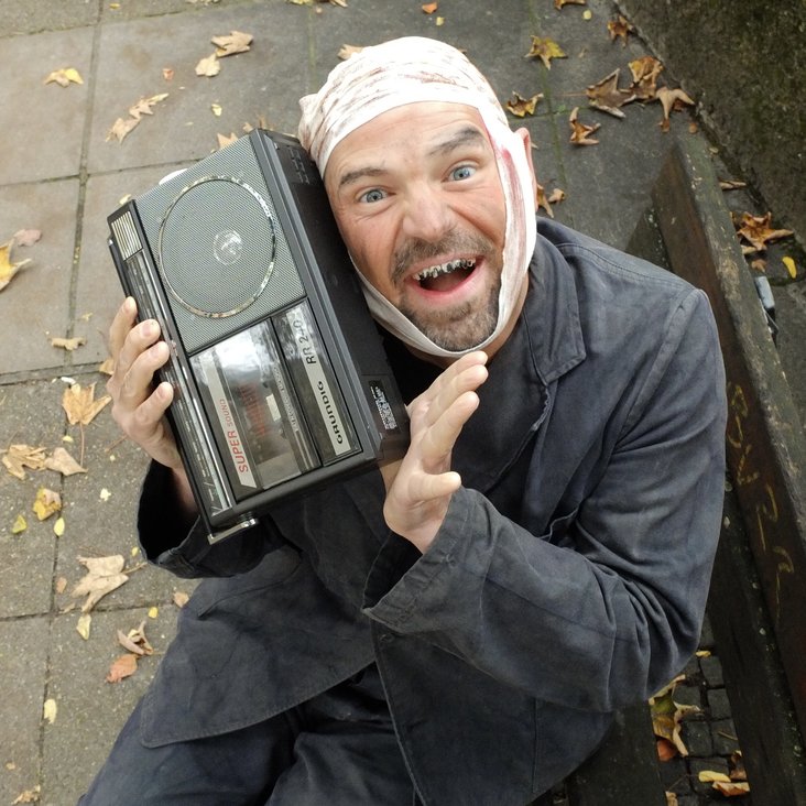 Ein Mann in schmutzigem Anzug, Kopfverband und schlechten Zähnen sitzt auf einer öffentlichen Parkbank und hat ein kleines Radio oder eine Jukebox in der Hand.