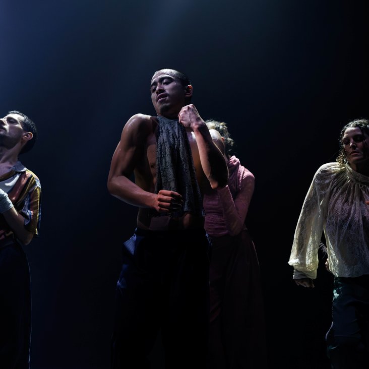 Die Tänzer:innen von Gauthier Dance in verschiedenfarbigen Kostümen auf der Bühne. Die Bühne ist auch dunkel und die Beleuchtung ist atmosphärisch.