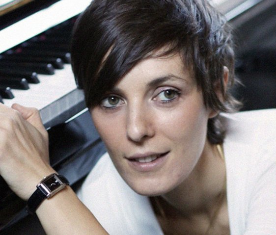 Porträt einer Komponistin am Klavier, Blick zur Kamera, ruhige Atelieratmosphäre.