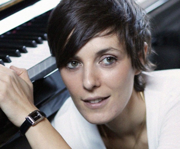 Porträt einer Komponistin am Klavier, Blick zur Kamera, ruhige Atelieratmosphäre.