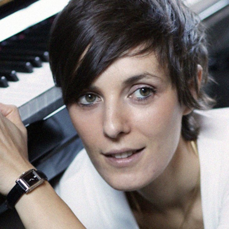 Porträt einer Komponistin am Klavier, Blick zur Kamera, ruhige Atelieratmosphäre.