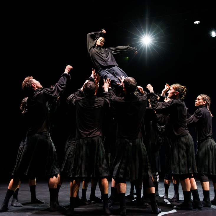 Die Tänzer:innen von Gauthier Dance in schwarzen Kostümen auf der Bühne. Die Bühne ist auch dunkel und es gibt wenig Licht.
