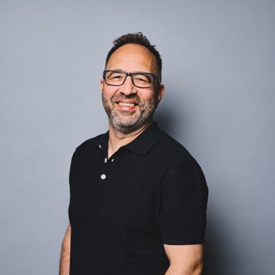 Portraitfoto von PIERO, er trägt eine blaue Jeans, ein schwarzes Poloshirt und eine Brille mit schwarzem Rahmen und blickt lächelnd in die Kamera.