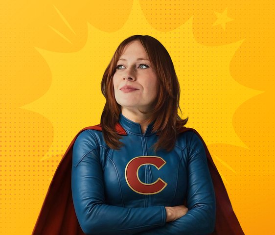 Die Künstlerin steht mit verschränkten Armen in einem Superheldinnenanzug mit Cape da und schaut links an der Kamera vorbei. Der Hintergrund ist gelb.