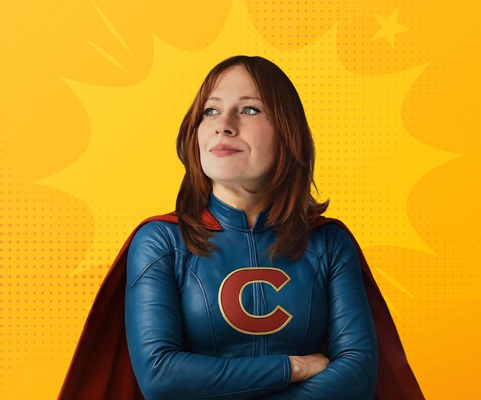 Die Künstlerin steht mit verschränkten Armen in einem Superheldinnenanzug mit Cape da und schaut links an der Kamera vorbei. Der Hintergrund ist gelb.