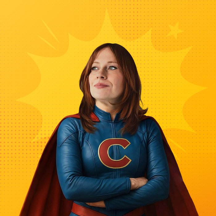 Die Künstlerin steht mit verschränkten Armen in einem Superheldinnenanzug mit Cape da und schaut links an der Kamera vorbei. Der Hintergrund ist gelb.