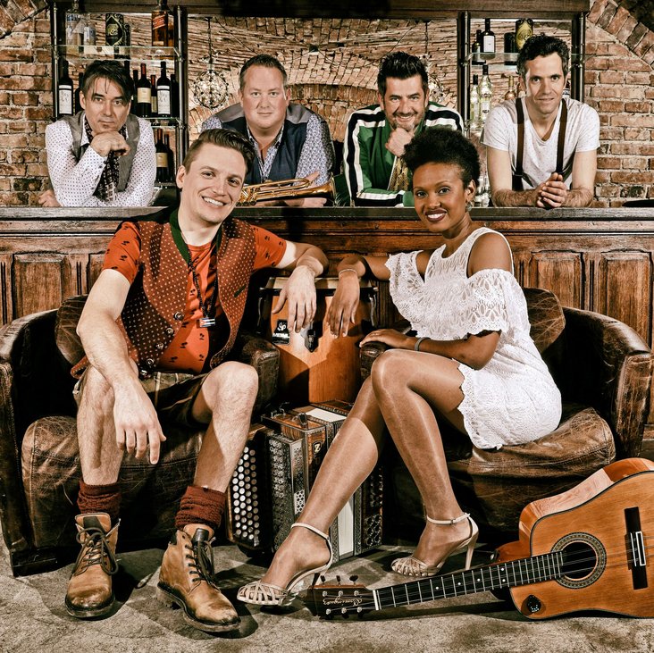 Zwei der Musiker:innen sitzen im Vordergrund auf einem Sofa, sie haben eine Gitarre und ein Akkordeon. Weiter hinten sind hinter einer Bar aus Holz vier weitere Musiker zu sehen.