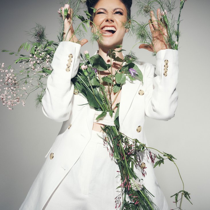 Portraitfoto von Tahnee in einem weißen Blazer mit verschiedenen Blüten und Blumen um sie und an ihr