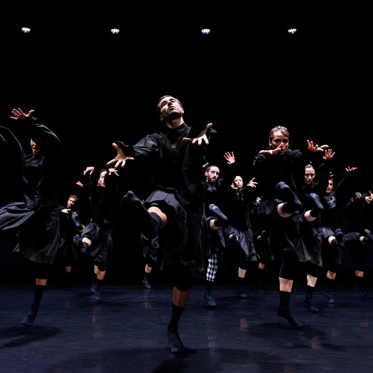 Die Tänzer:innen von Gauthier Dance in schwarzen Kostümen auf der Bühne. Die Bühne ist auch dunkel und es gibt wenig Licht.