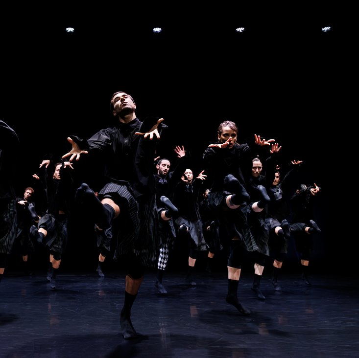 Die Tänzer:innen von Gauthier Dance in schwarzen Kostümen auf der Bühne. Die Bühne ist auch dunkel und es gibt wenig Licht.