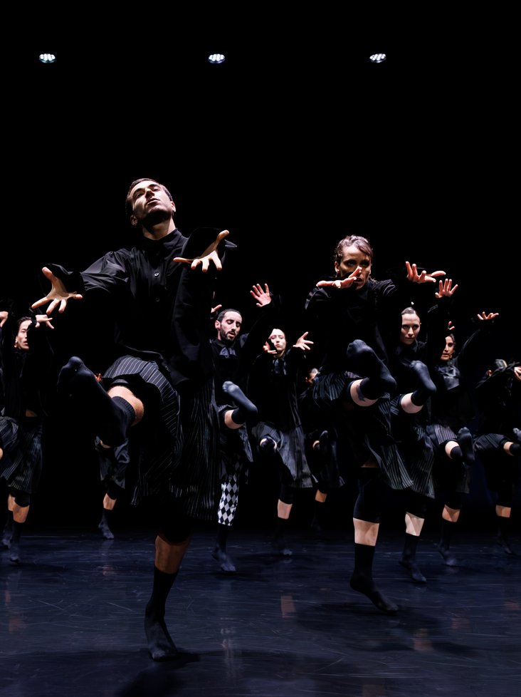 Die Tänzer:innen von Gauthier Dance in schwarzen Kostümen auf der Bühne. Die Bühne ist auch dunkel und es gibt wenig Licht.