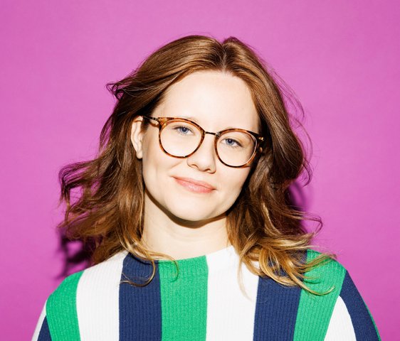 Leonie Schöler trägt eine Brille und einen längs gestreiften Pullover in den Farben weiß, blau und grün. Sie steht vor einer lilafarbenen Wand.