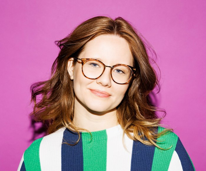 Leonie Schöler trägt eine Brille und einen längs gestreiften Pullover in den Farben weiß, blau und grün. Sie steht vor einer lilafarbenen Wand.