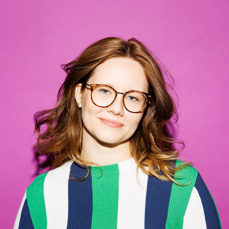 Leonie Schöler trägt eine Brille und einen längs gestreiften Pullover in den Farben weiß, blau und grün. Sie steht vor einer lilafarbenen Wand.