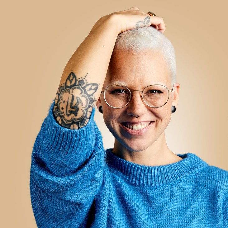Stefanie Heinzmann schaut lächelnd in die Kamera. Sie trägt eine helle Brille, schwarze Ohrringe, einen blauen Pullover, bei welchem sie die Ärmel hochgekrempelt hat und ihre kurzen Haare sind hellblond. Ihren rechten Arm hat sie angehoben, sodass ihre Hand auf ihrem Kopf aufliegt und der Ellenbogen in Richtung der Kamera zeigt. An ihrem rechten Ellenbogen hat sie ein Ornament-Tattoo.