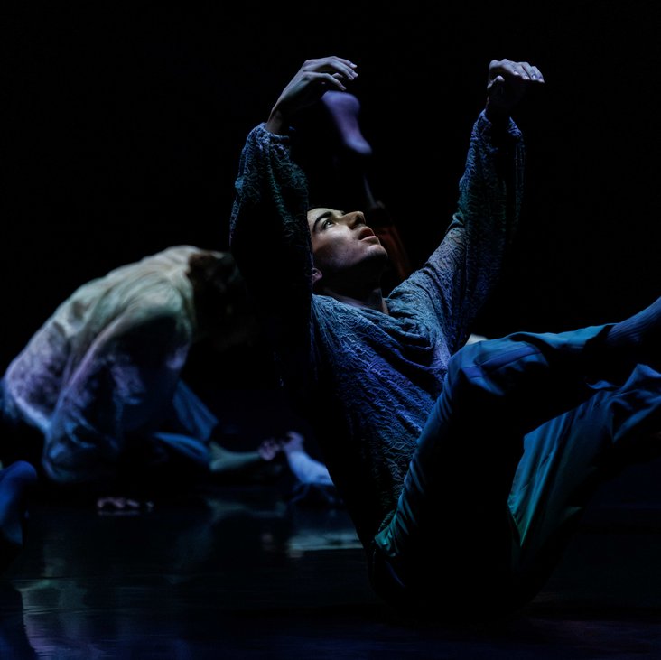 Die Tänzer:innen von Gauthier Dance in verschiedenfarbigen Kostümen auf der Bühne. Die Bühne ist auch dunkel und die Beleuchtung ist atmosphärisch.
