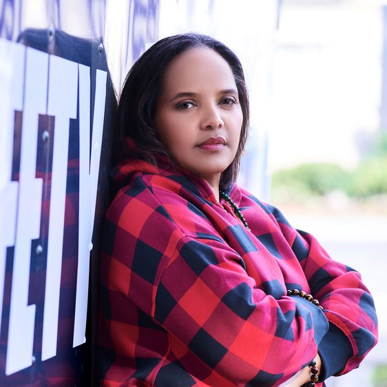 Portraitfoto von Terri Lyne Carrington. Sie lehnt an einer Wand mit Aufdruck, trägt ein schwarz-rotes Oberteil mit großen Karos und hat die Arme verschränkt. Sie blickt nach rechts in die Kamera.