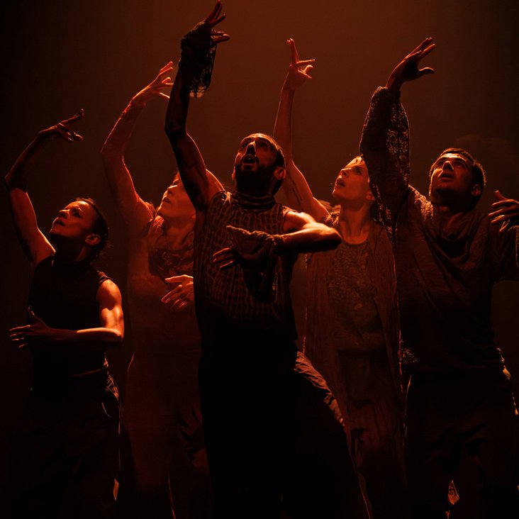 Die Tänzer:innen von Gauthier Dance in verschiedenfarbigen Kostümen auf der Bühne. Die Bühne ist auch dunkel und die Beleuchtung ist atmosphärisch.