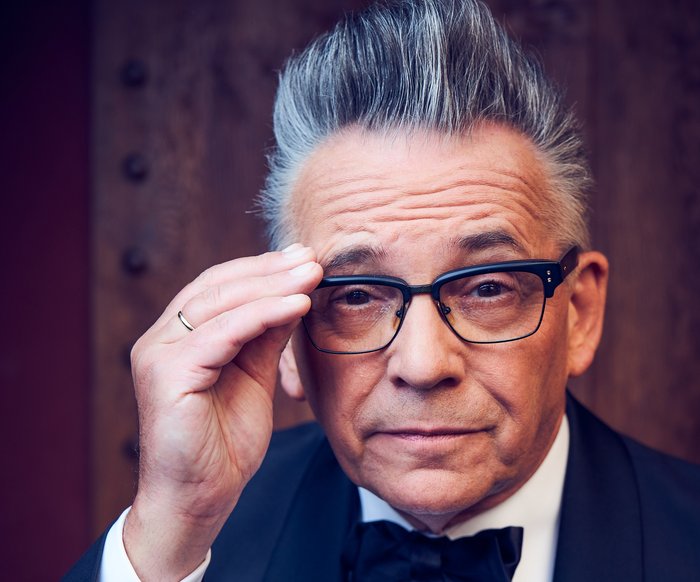 Ein Mann im Smoking blickt in die Kamera. Er trägt eine Brille, die er mit einer Hand festhält. Seine Haare sind grau und toupiert, er zieht eine Augenbraue hoch.