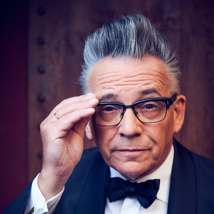 Ein Mann im Smoking blickt in die Kamera. Er trägt eine Brille, die er mit einer Hand festhält. Seine Haare sind grau und toupiert, er zieht eine Augenbraue hoch.