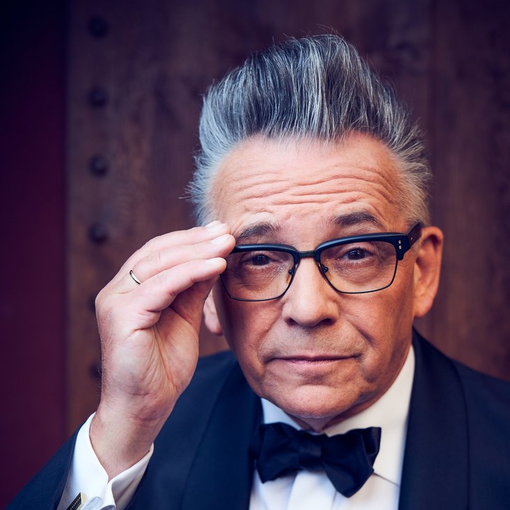 Ein Mann im Smoking blickt in die Kamera. Er trägt eine Brille, die er mit einer Hand festhält. Seine Haare sind grau und toupiert, er zieht eine Augenbraue hoch.