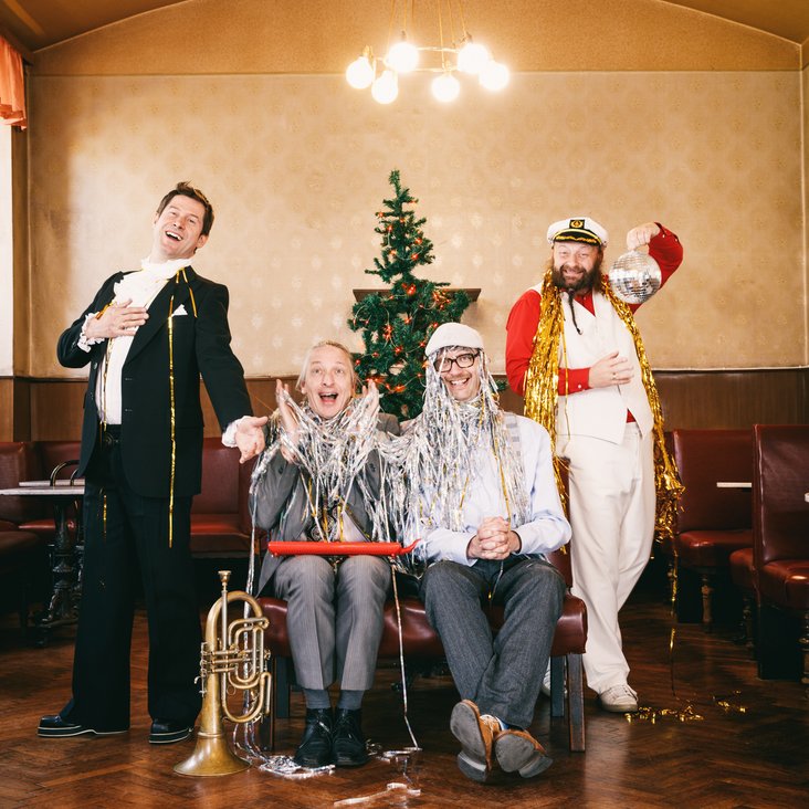 Die Künstler sitzen und stehen vor einem Weihnachtsbaum, sie selbst sind teilweise mit Lametta geschmückt