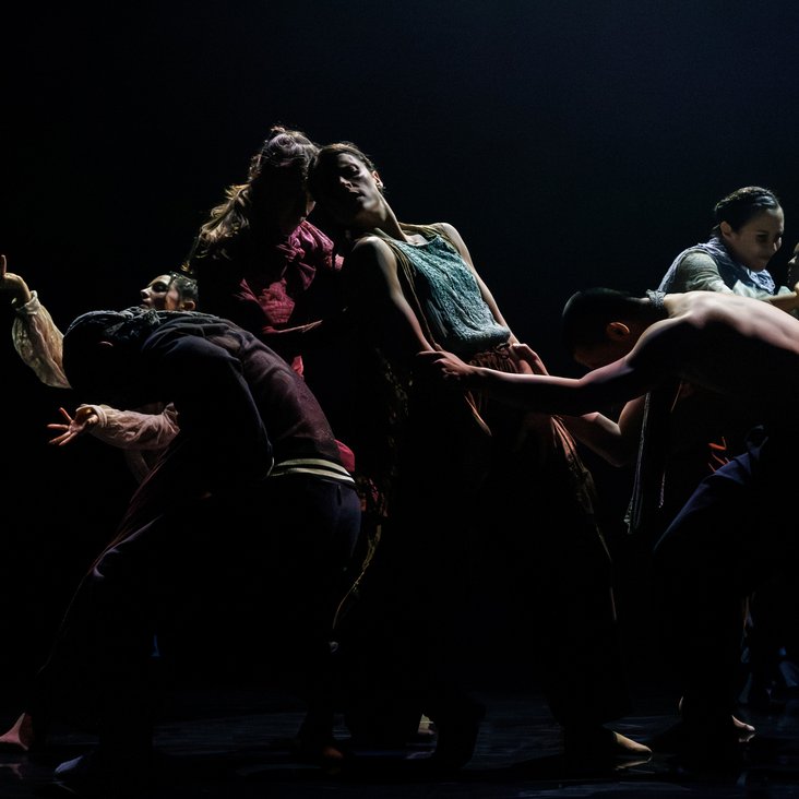 Die Tänzer:innen von Gauthier Dance in verschiedenfarbigen Kostümen auf der Bühne. Die Bühne ist auch dunkel und die Beleuchtung ist atmosphärisch.