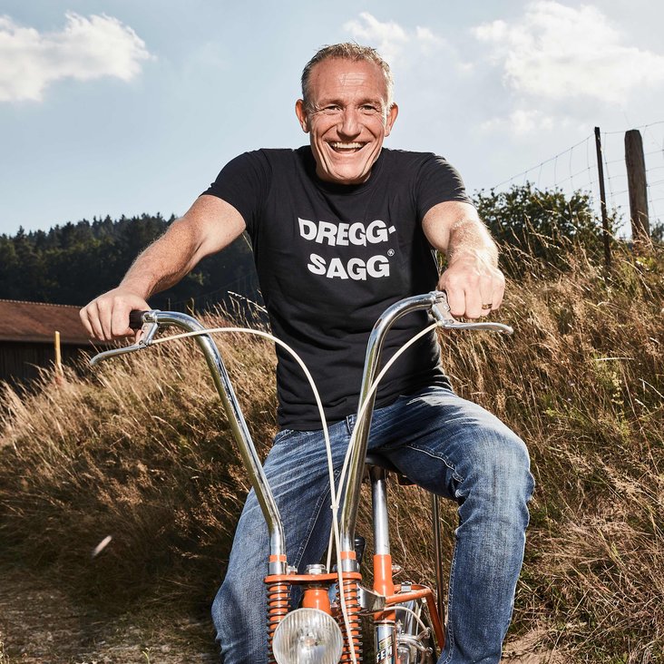 Michl Müller sitzt auf einem orangefarbenen Fahrrad, er fährt auf einer Wiese oder einem Feld Richtung Fotograf:in. Er trägt Jeans und ein schwarzes T-Shirt mit der Aufschrift "Dregg-Sagg"