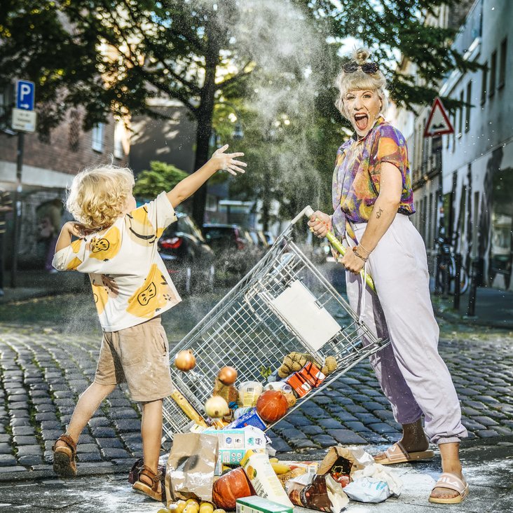 Eine Frau und ein Kind stehen auf einem Gehweg. Das Kind wirft mit Mehl, die Frau hält einen umgefallenen Einkaufswagen, aus dem die Einkäufe hinausfallen.  Die Frau schaut in die Kamera und schreit.