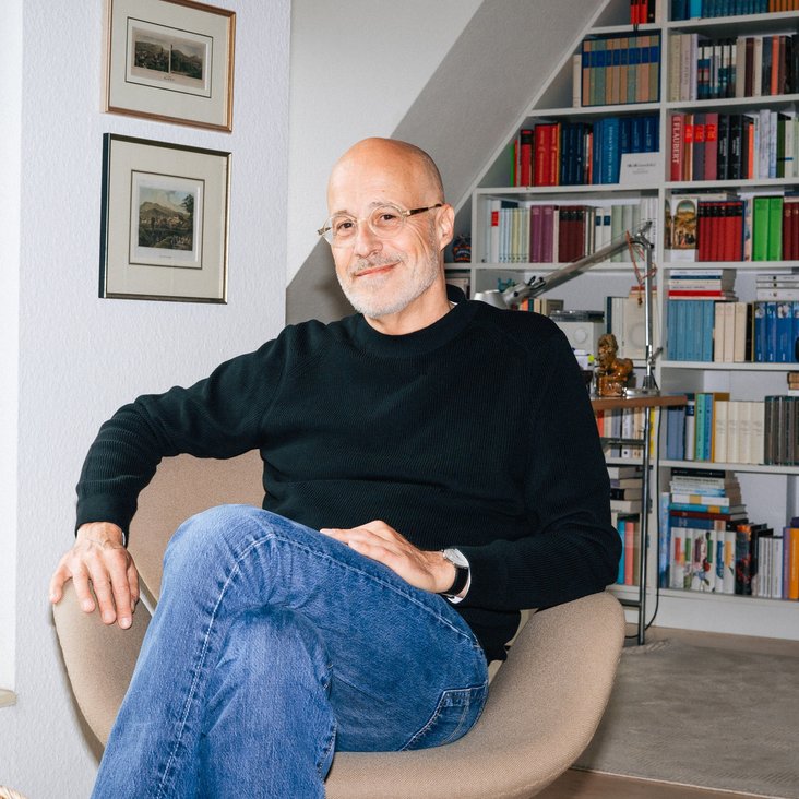 Portraitfoto von Daniel Haas. Er trägt Jeans und einen schwarzen Pullover und sitzt in einem Sessel. Im Hintergrund sind Bücherregale zu sehen.