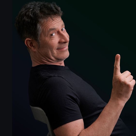 Rolf Miller sitzt auf einem Stuhl. Er trägt ein schwarzes T-Shirt, der Hintergrund ist dunkel. Er sitzt seitlich zur Kamera, dreht den Kopf und hebt die rechte Hand mit erhobenem Zeigefinger.